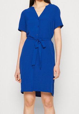Femme portant une robe bleue à manches courtes, longueur genoux, avec des boutons et une ceinture nouée à la taille, debout devant un fond uni.
