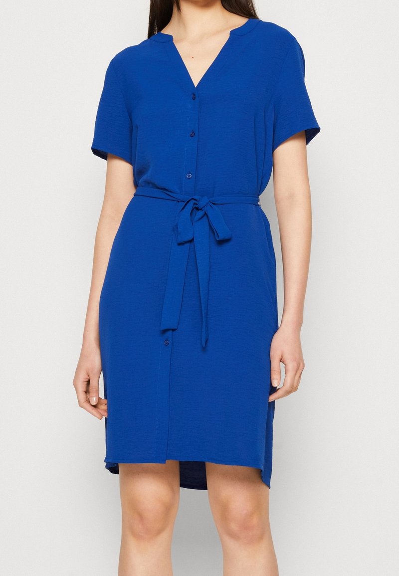 Femme portant une robe bleue à manches courtes, longueur genoux, avec des boutons et une ceinture nouée à la taille, debout devant un fond uni.
