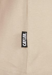 Beige stof met een gladde textuur, voorzien van een zwart label met witte tekst "CAYLER." Het kledingstuk heeft nette stiksels en een eenvoudig ontwerp.