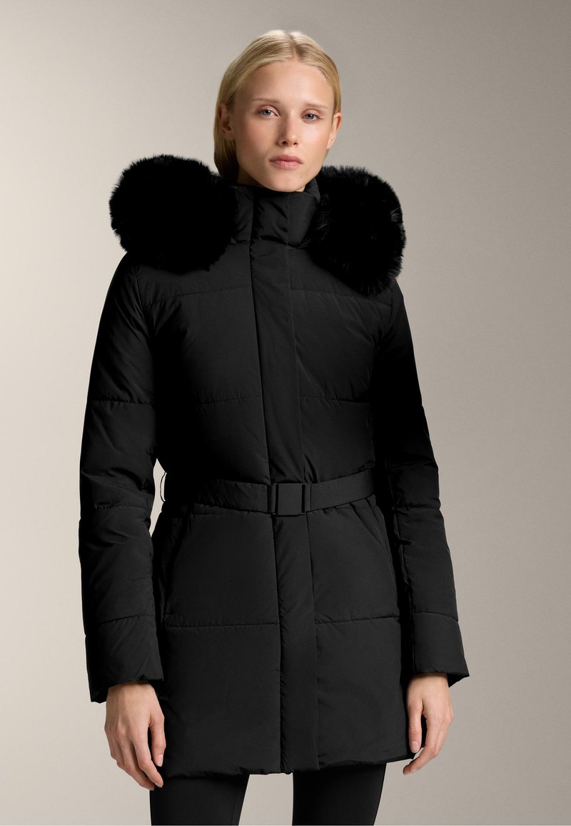 Svart puffjacka med bälte i midjan och en stor, fluffig svart pälskrage. Har en quiltad textur och långa ärmar.