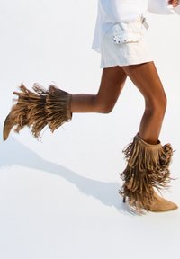 Botki z frędzlami w kolorze jasnobrązowym zamszowe, z noskiem w szpic, ozdobione warstwowymi frędzlami wzdłuż cholewki, które tworzą teksturę i ruch.