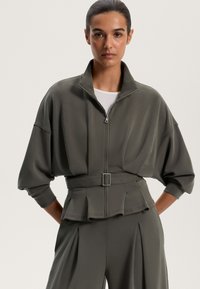 Olivno zelena jakna z zadrgo in visokim ovratnikom, prevelikimi rokavi in pasom na pasu. Gladek material z nabranimi detajli na robu.