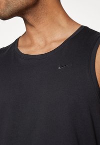 Débardeur Nike noir avec un col rond et sans manches. Présente un petit logo swoosh noir sur la poitrine supérieure gauche. Texture douce et lisse.