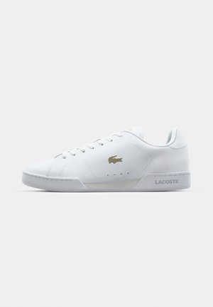 Bílé tenisky Lacoste, hladký syntetický materiál, kulatá špička, šněrování na sedmi dírkách, zlatý logový akcent na boku, texturovaná gumová podrážka, plochý podpatek.