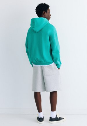 Uomo che indossa una felpa con cappuccio color teal, pantaloncini grigio chiaro fino al ginocchio, calzini bianchi e scarpe da ginnastica nere, in piedi di fronte a un muro bianco.