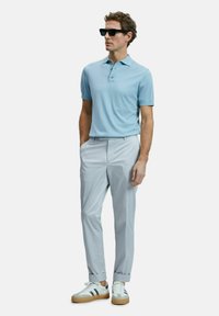 Helles blaues Poloshirt mit kurzen Ärmeln, hellgraue Anzughose mit hochgeschlagenen Säumen und weiße Sneaker mit schwarzen Akzenten. Schwarze Sonnenbrille.