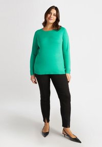 Suéter verde de manga larga con cuello redondo, puños acanalados y dobladillo. Combinado con pantalones negros ajustados con aberturas en los tobillos.
