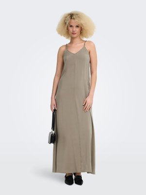 Vrouw met krullend blond haar, gekleed in een lange taupekleurige slipjurk, met een zwarte handtas, staand tegen een effen lichte achtergrond.