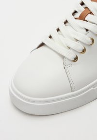 White leather sneaker with a rounded toe, textured rubber sole, gold eyelets, and cream laces, featuring a brown lace accent.