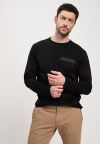 Czarna koszulka z długim rękawem z kieszenią na piersi z metką; gładki materiał i luźny krój. Model nosi beżowe spodnie.