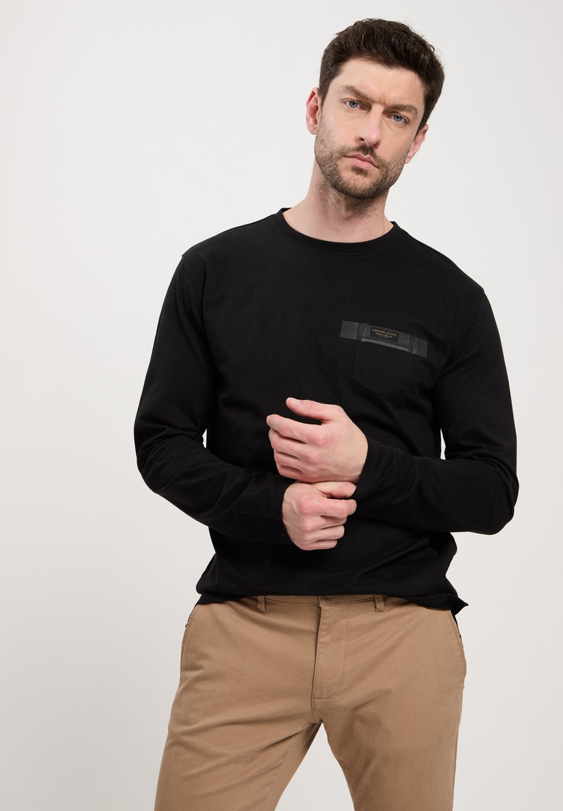 Czarna koszulka z długim rękawem z kieszenią na piersi z metką; gładki materiał i luźny krój. Model nosi beżowe spodnie.