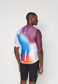 Maillot de cyclisme à manches courtes dans un dégradé de violet, bleu et orange. Il présente un tissu en maille, des poches au dos et le logo "LE COL".
