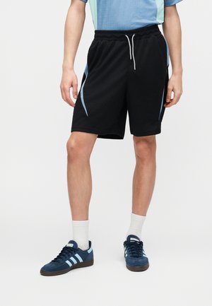 Person iført sorte træningsshorts med lyseblå sidelister, hvide sokker, blå sneakers og en lyseblå kortærmet skjorte.