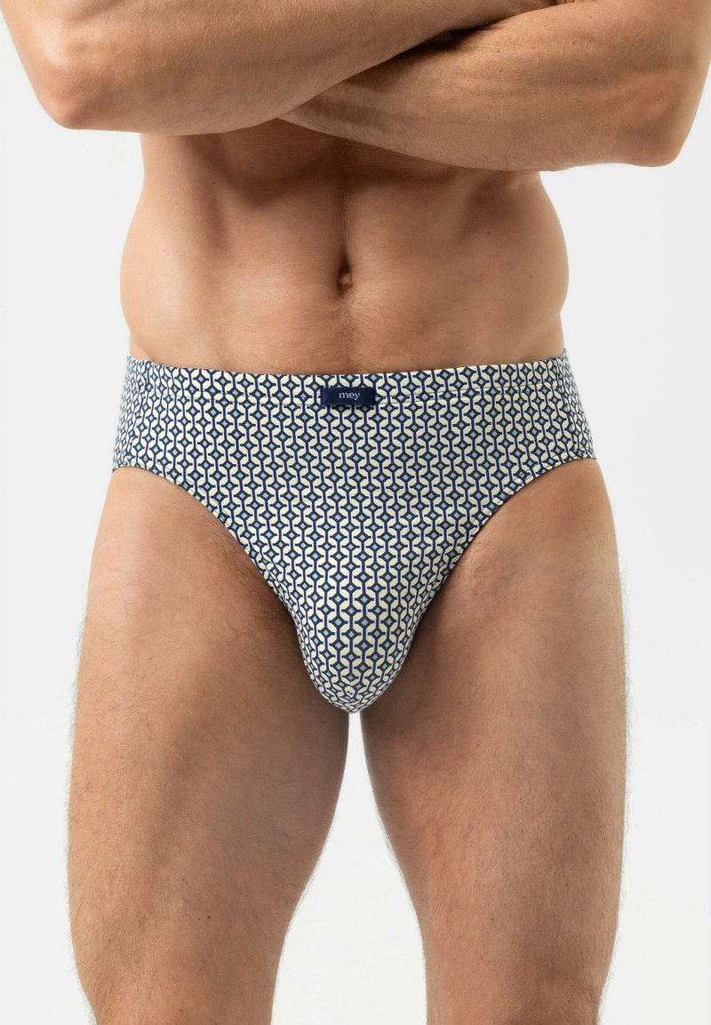 Torso masculin et parties supérieures des jambes portant un slip blanc avec un motif géométrique bleu, debout les bras croisés sur la poitrine.