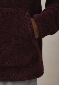 Vīnsarkans flīsa džemperis ar kabatu un teksturētu audumu. Rāda mīkstu, pūkainu virsmu un aproces uz piedurknēm ērtībai.