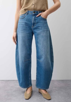 Persoon draagt blauwe jeans met wijde pijpen, beige mouwloze top en lichtbruine instapschoenen, staand tegen een effen lichte achtergrond.