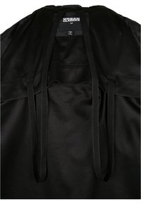 Vêtement en tissu mesh noir avec étiquette visible indiquant Urban Classics M Made in China, et deux cordons noirs suspendus.