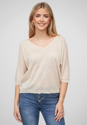 Lightweight, beige knit sweater with a V-neck, half sleeves, and a chevron pattern. Paired with blue denim jeans.