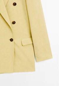 Cremefarbener Blazer aus strukturierter Stoff, mit drei dunklen Knöpfen und einer einzelnen Tasche auf der linken Seite.