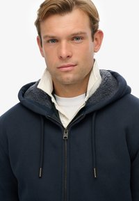 Marineblå zip-up hoodie med en kontrasterende grå fleece-foderet hætte, med lynlås og snore. Indersiden af kraven har en hvid skjorte nedenunder.