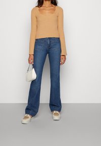 Beige rippad långärmad topp med fyrkantig halsringning, tillsammans med blå utsvängda jeans. Vit handväska med texturerad yta och sneakers.