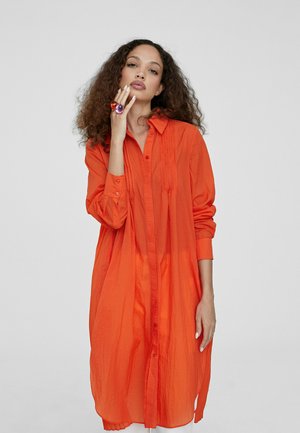 Vestido camisa naranja con un frente de botones, mangas largas y tela texturizada. Acentuado con un anillo morado y naranja en la mano.
