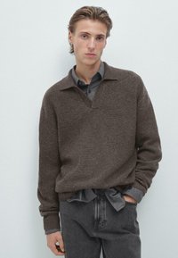 Pull en tricot marron avec un col en V et une texture côtelée, superposé sur une chemise gris à boutons. Jean slim-fit gris foncé.