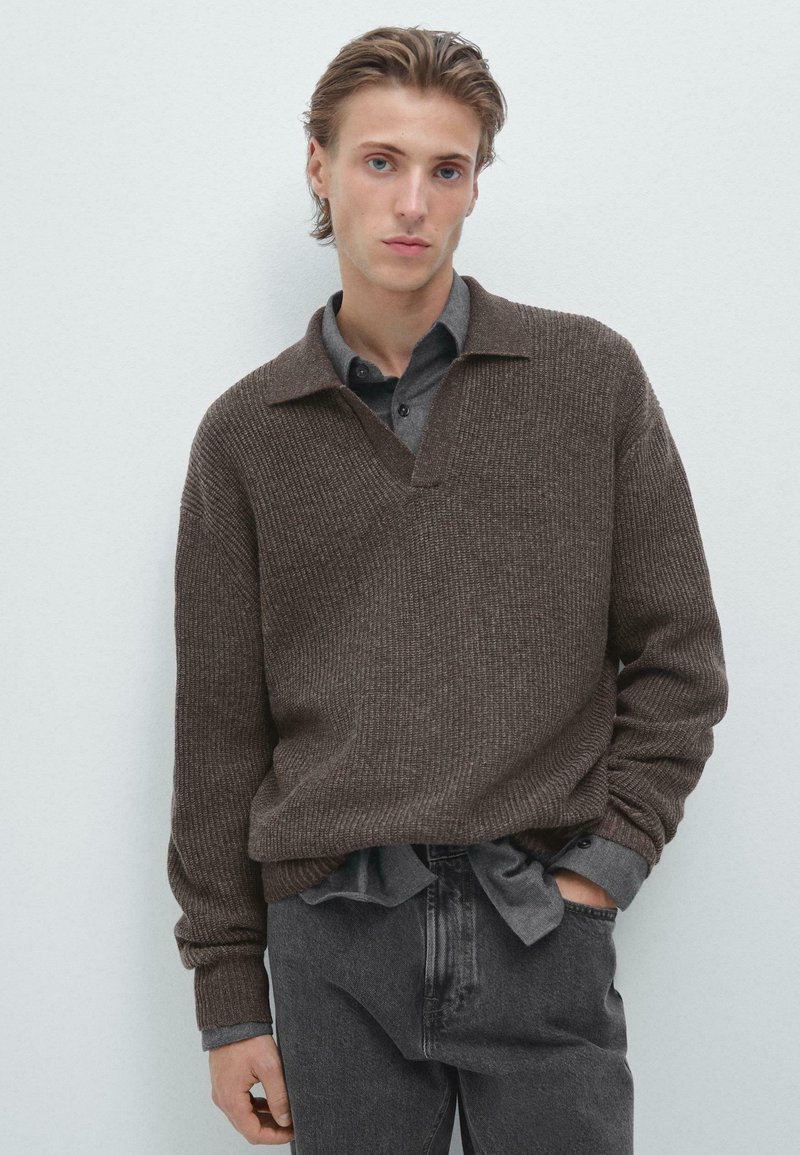 Pull en tricot marron avec un col en V et une texture côtelée, superposé sur une chemise gris à boutons. Jean slim-fit gris foncé.