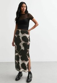 Top corto de malla negra de manga corta combinado con una falda larga y estampada que presenta tonos verde oscuro y beige. La falda tiene aberturas laterales y se lleva con zapatos negros.
