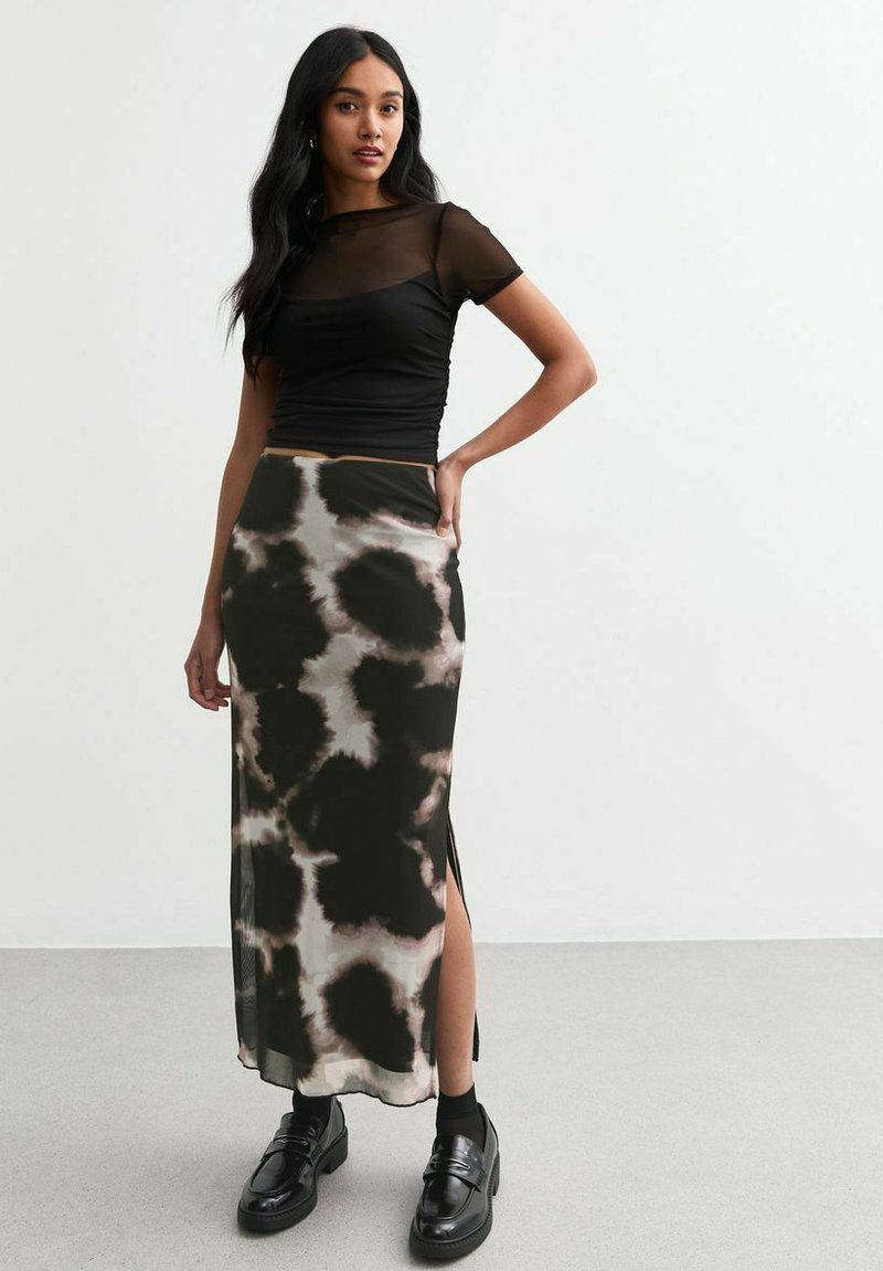 Top corto de malla negra de manga corta combinado con una falda larga y estampada que presenta tonos verde oscuro y beige. La falda tiene aberturas laterales y se lleva con zapatos negros.