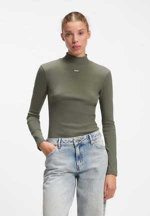 Donna che indossa una maglia aderente verde oliva a maniche lunghe e jeans azzurri a vita alta, in piedi con una mano in tasca su sfondo bianco.