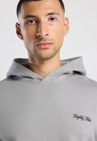 Giovane uomo con capelli corti e scuri che indossa un hoodie grigio chiaro con la scritta "Eighty Nine" ricamata in scuro sul petto.