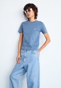 Helles blaues T-Shirt mit kurzen Ärmeln und Rundhalsausschnitt, versehen mit kleinem Logo, kombiniert mit lockeren, hellblauen Jeans. Silberne, verspiegelte Sonnenbrille.