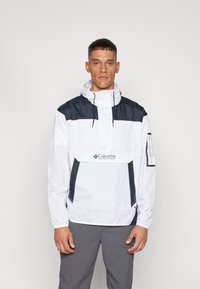 Jachetă albă rezistentă la apă cu accente bleumarin, glugă și buzunar frontal. Are manșete elastice și branding pe piept. Material ușor.