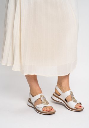 Feet wearing white sandals with decorative pearls and beads, paired with a cream pleated skirt against a plain background.