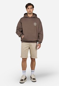 Brun hættetrøje med frontlomme, broderet logo, parret med beige shorts og grå sneakers. Model står foran en simpel hvid baggrund.
