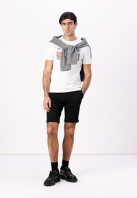 Camiseta blanca con diseño gráfico, pantalones cortos negros, suéter gris drapeado sobre los hombros y zapatos negros, con cabello texturizado y calcetines.