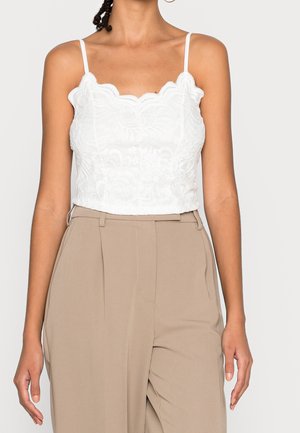 Mujer vistiendo un top corto de encaje blanco con tirantes finos y pantalones beige de talle alto sobre un fondo liso.