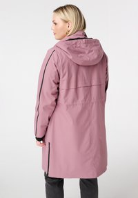 Vrouw die zijwaarts kijkt, gekleed in een rozenkleurige regenjas met capuchon en zwarte accenten, met donkergrijze broek, op een witte achtergrond.