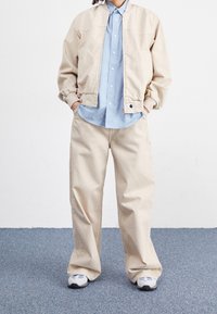 Veste bomber beige portée sur une chemise à boutons bleu clair, associée à un pantalon beige ample et des baskets grises. Design simple et coupe décontractée.