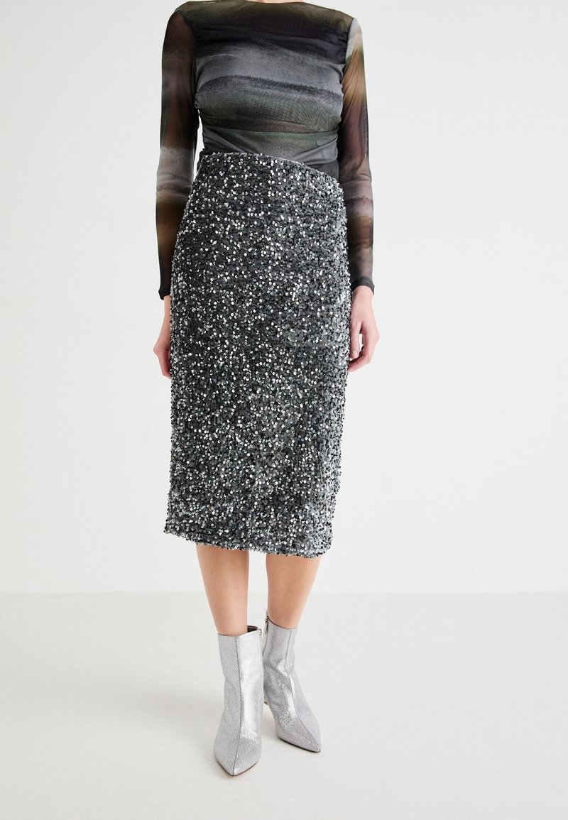 Jupe noire à paillettes longueur genou associée à des bottines argentées et un haut noir et gris transparent à manches longues sur un mannequin.