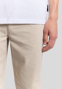 Pantaloni di cotone beige con una vestibilità rilassata, dotati di tasche laterali e cuciture a vista. Una maglietta bianca è parzialmente visibile sopra.