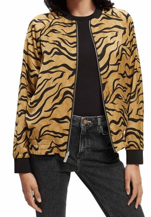 Frau trägt eine beige-schwarze, tigergestreifte Zip-Up-Jacke, schwarzes Shirt und hoch taillierte schwarze Jeans, steht mit entspannt seitlich hängenden Händen.