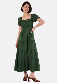 Grünes Baumwoll-Maxikleid mit Puffärmeln, quadratischem Ausschnitt, gerafftem Oberteil und gestuften Rock. Getragen mit braunen Blockabsatzsandalen.