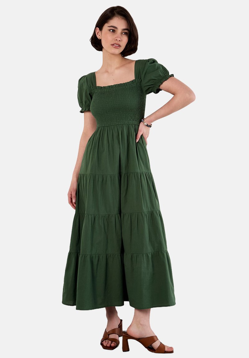 Grünes Baumwoll-Maxikleid mit Puffärmeln, quadratischem Ausschnitt, gerafftem Oberteil und gestuften Rock. Getragen mit braunen Blockabsatzsandalen.