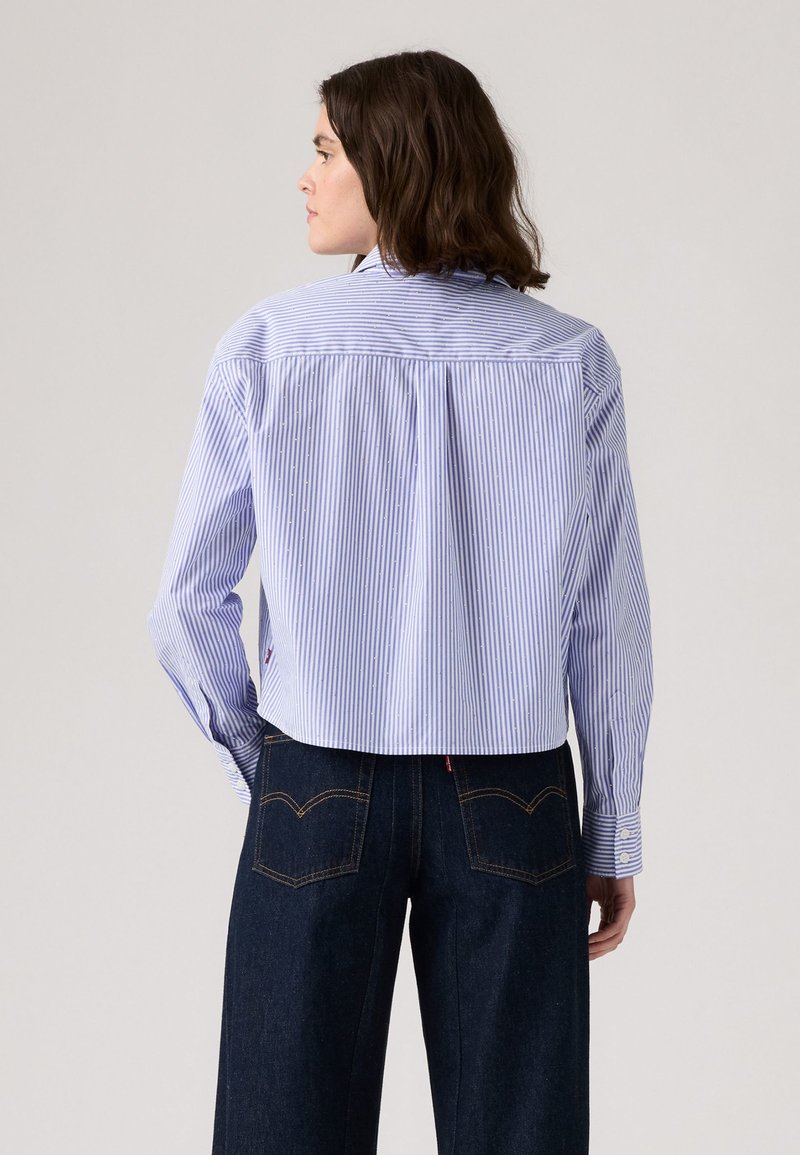 Chemise à rayures, bleu clair, à boutons, avec des manches longues, au design court, et des plis dans le dos. Associée à un pantalon en denim foncé avec des poches arrière.