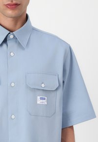 Lys blå kortermet skjorte laget av teksturert stoff, med krage som kan kneppes, to brystlommer og et merke ved lommen.