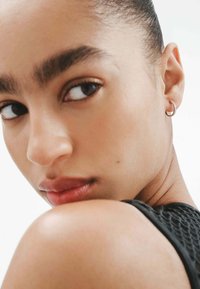 Primer plano de la cara de un modelo, mostrando piel suave, cejas definidas y maquillaje sutil. Un pequeño pendiente de aro plateado complementa el look.