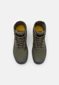 Bottines kaki avec capuchons en caoutchouc noir, tige en tissu texturé, dotées d'un laçage et d'une doublure intérieure jaune avec une marque.