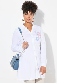 Weißes Langarmhemd mit Blumenmotiven in Blau und Pink, kombiniert mit einer hellblauen Handtasche mit goldener Kettenriemen.